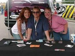 Three people at a radio station remote broadcast table. Woman on left has long red hair, man in the middle is a blonde with sunglasses on and woman on the right is blonde, wearing a pink blouse.