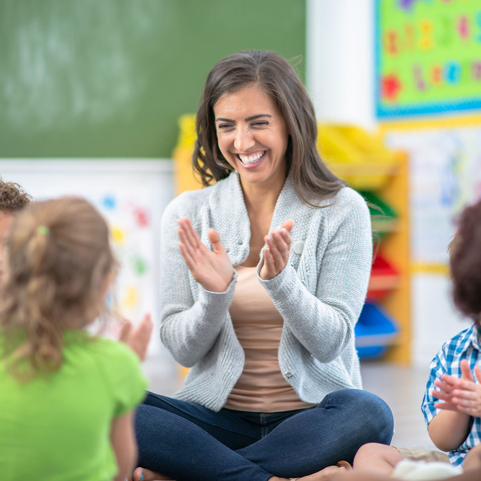 Teacher claps and smiles with young children in a colorful classroom. Kids sit on floor, engaged. Atmosphere is joyful and playful.