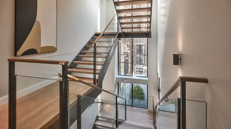 Multi-level NYC townhouse stair with glass railings, warm wood treads, and tall windows