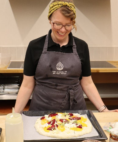 LIANNA COURT, Food Education Specialist in a kitchen wearing an apron smiles at an uncooked pizza with colorful toppings on a tray.