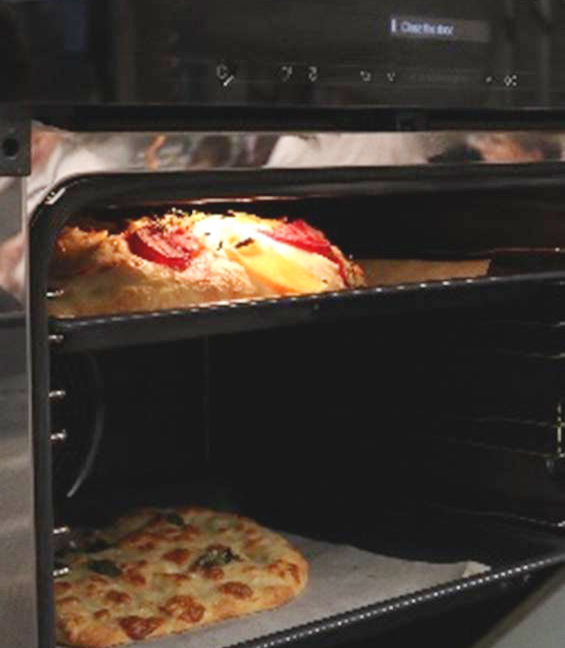Pizzas bake in an oven with one on a rack and another below on a tray.