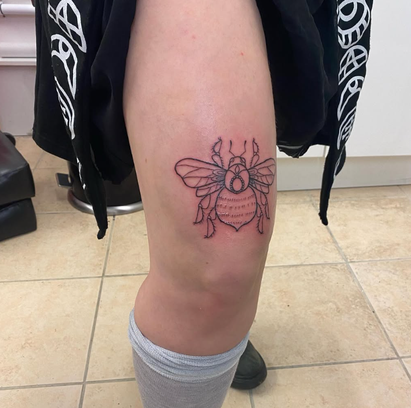 A bee tattoo on a person's thigh in a bright room. The design is black outline, and they wear a gray sock and black clothing with white patterns.