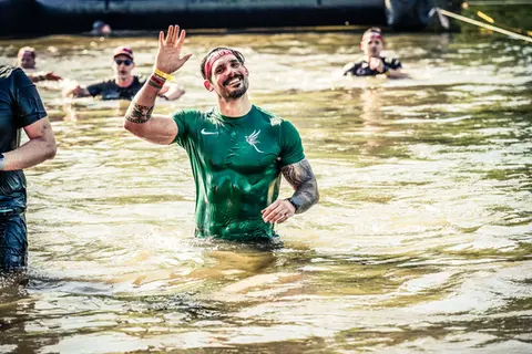 Mann winkt im schlammigen Wasser, grünes Shirt, Hindernislauf
