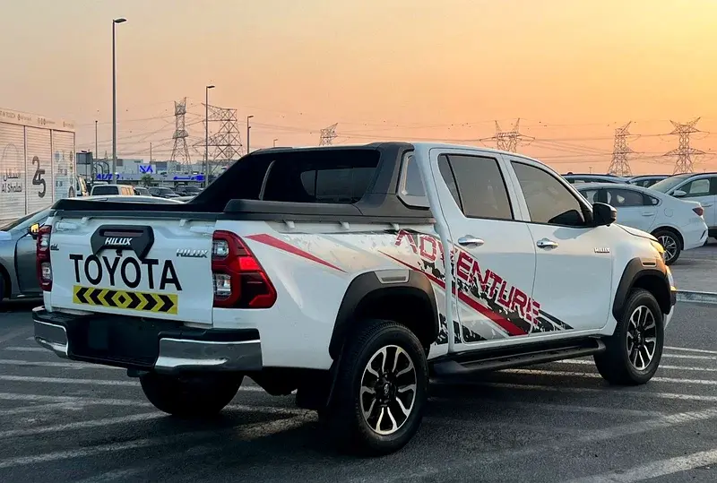Miniature : Camionnette Toyota Hilux blanche avec autocollant ADVENTURE, stationnée au coucher du soleil.