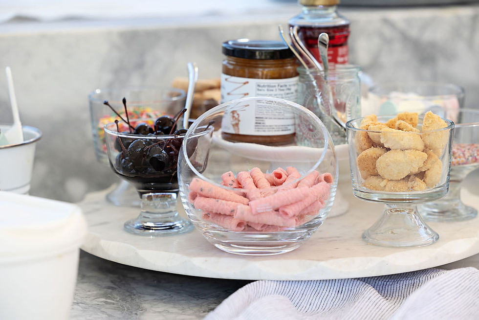 Assorted ice cream toppings displayed in glass bowls, including maraschino cherries, rainbow sprinkles, pink rolled wafers, and cookies on a marble serving tray.