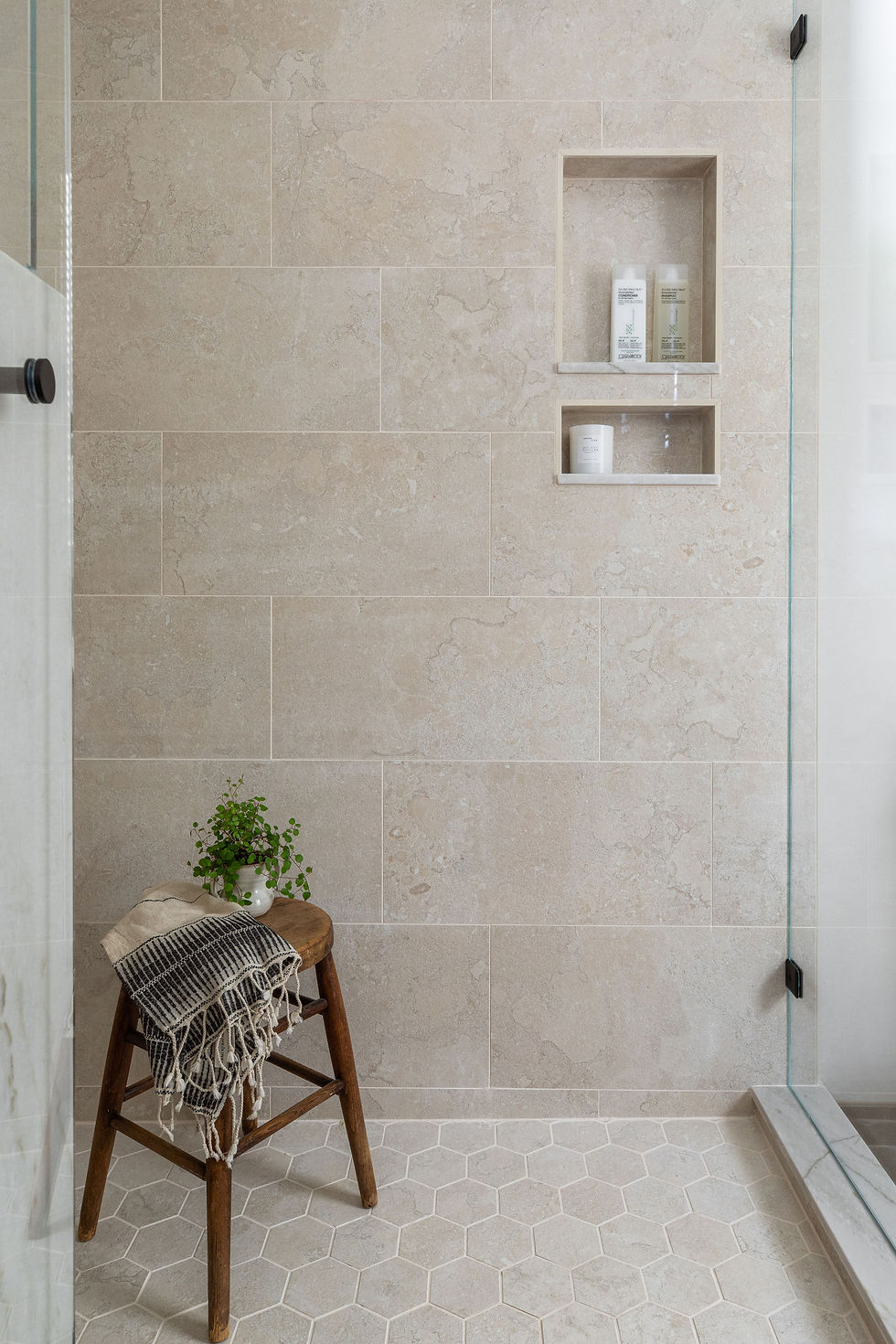 Neutral-toned shower with warm beige stone tiles in a stacked layout, paired with a light hex tile floor. Two recessed niches provide functional storage, styled simply with bath essentials, while a rustic wooden stool and greenery bring an inviting, organic touch.