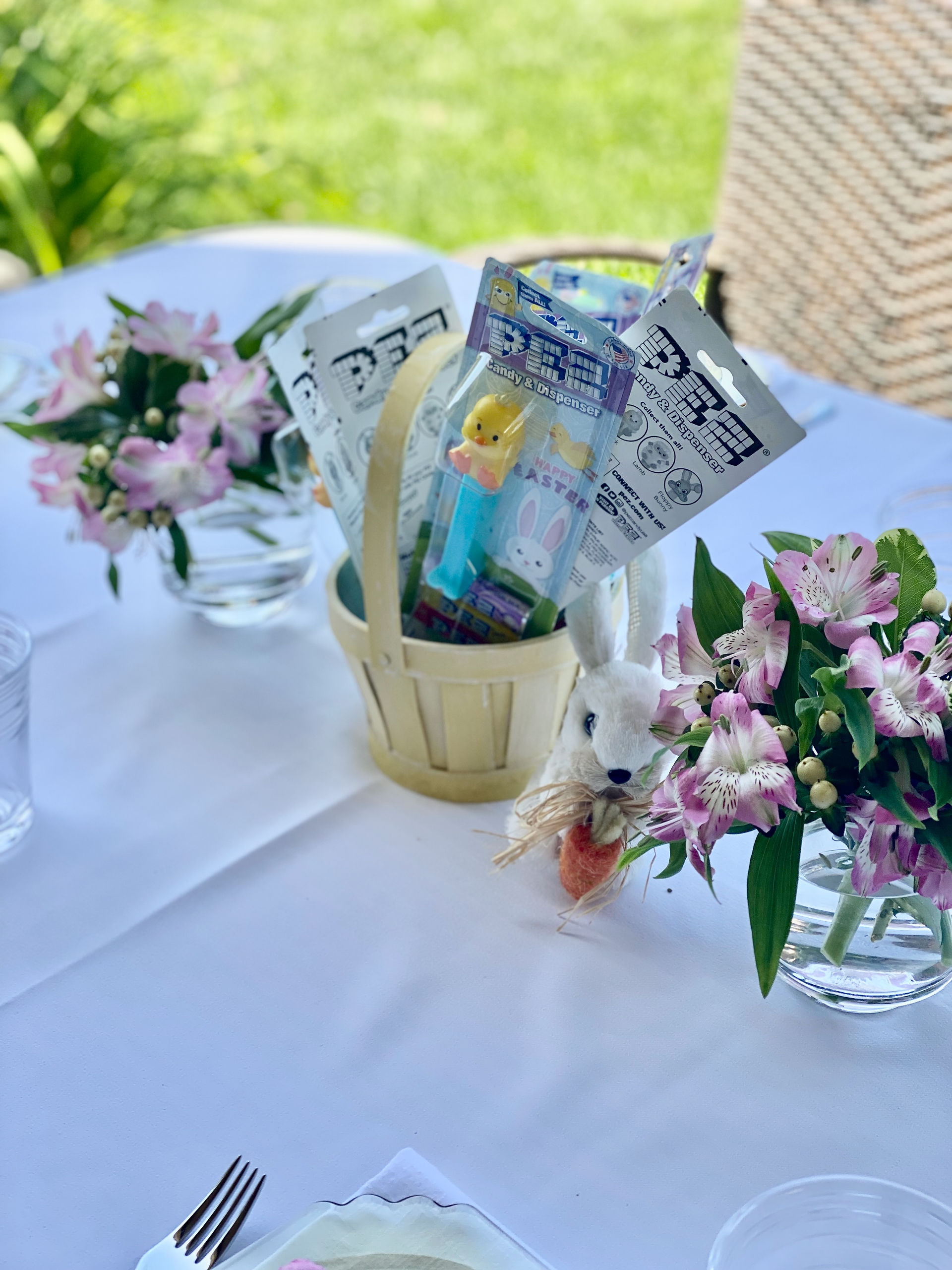 Kids' Easter table centerpiece featuring a yellow basket filled with PEZ candy dispensers, pastel flowers, and a small bunny figurine, set on a white tablecloth