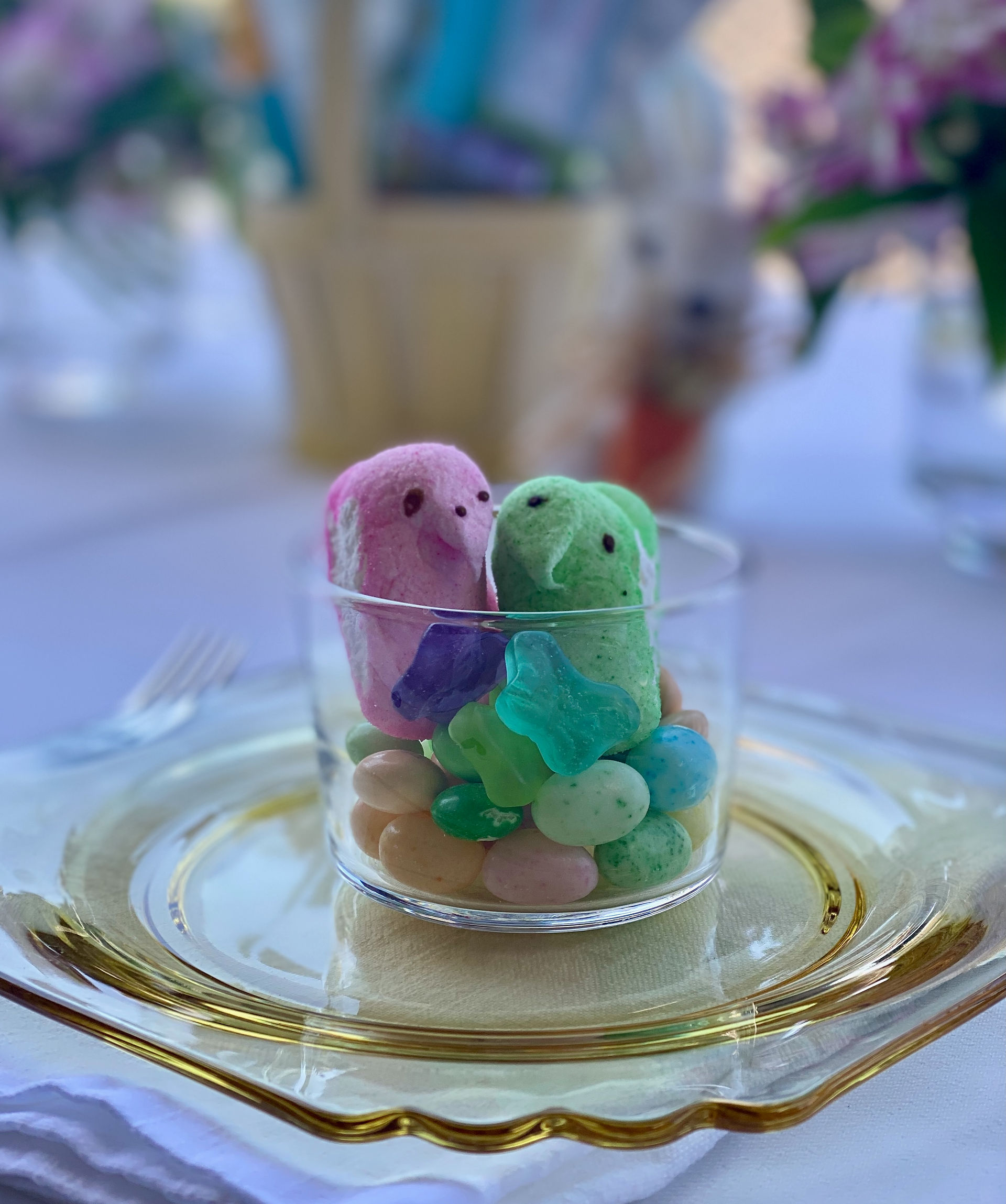 Clear glass cup filled with pastel Easter candy and colorful marshmallow Peeps, set on a gold-rimmed plate as a festive treat at a kids' place setting