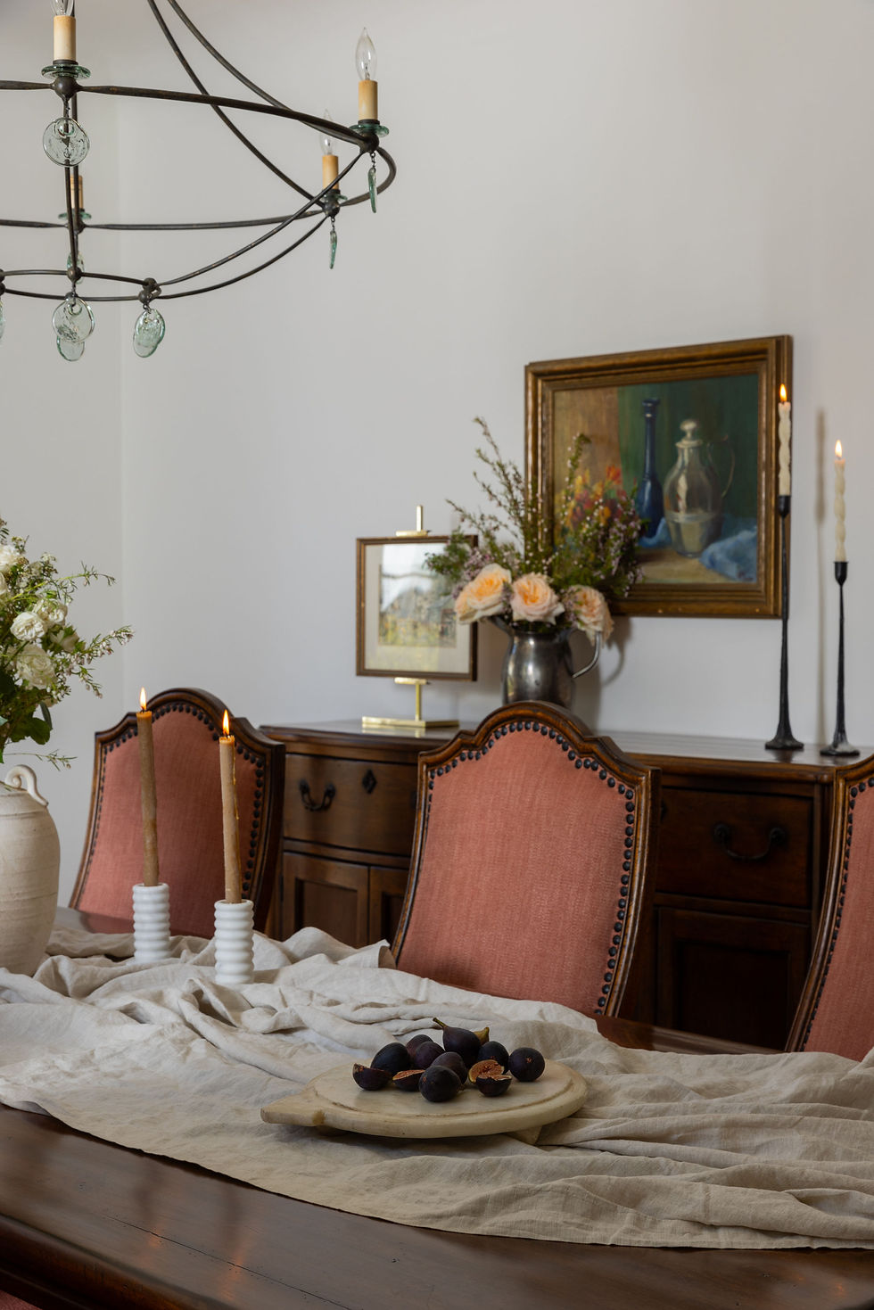 Elegant dining room with antique wood furniture, terracotta upholstered chairs, soft linen table runner, and curated still-life artwork creating a layered, lived-in European charm.