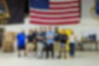 Group of nine people smiling in a workshop with an American flag, surrounded by boxes and equipment. Casual attire, collaborative mood.