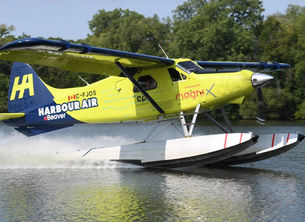 Bright yellow-and-blue Harbour Air eBeaver floatplane C-FJOS skims across a lake, spraying water beside green trees.
