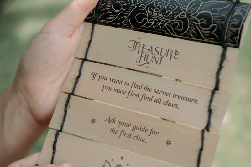 Child hands hold a treasure hunt scroll lontar, displaying clues and decorative patterns of niskala tours. The background is blurred green grass.