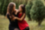 Woman and girl in a christmas tree farm share a joyful moment, smiling at each other. The girl wears a red dress; trees and grass fill the background.