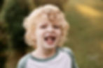 A smiling child with curly blonde hair in a green and white shirt, outdoors with blurred greenery. Christmas tree farm