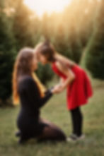 Woman and girl in dresses share a joyful forehead touch in a sunny field with christmas trees. Girl wears red, woman in black. Warm, loving scene.