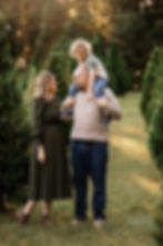 A family in fall attire, with a child on a man's shoulders, stands among evergreen trees. Warm sunlight filters through the trees, creating a cozy mood.