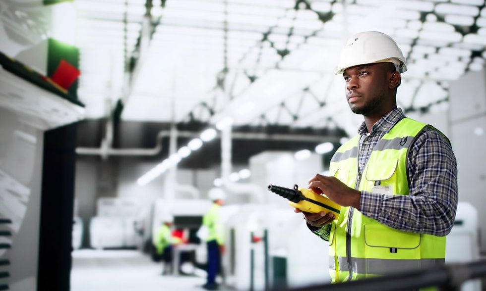 A worker wearing a high-visibility vest and hard hat inspects a machine in a warehouse with other workers behind them.