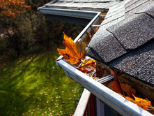 Orange and brown autumn leaves filling a metal rain gutter along a dark shingled roof with trees in the background.