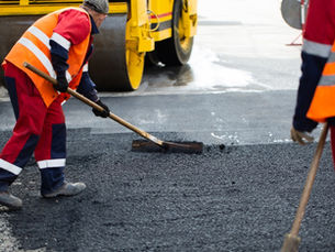 Road workers in orange safety vests smoothing fresh asphalt on a street beside a yellow road roller.