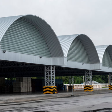 A row of large warehouse bays with curved metal roofs, striped pillars, stacked pallets, and wet pavement.