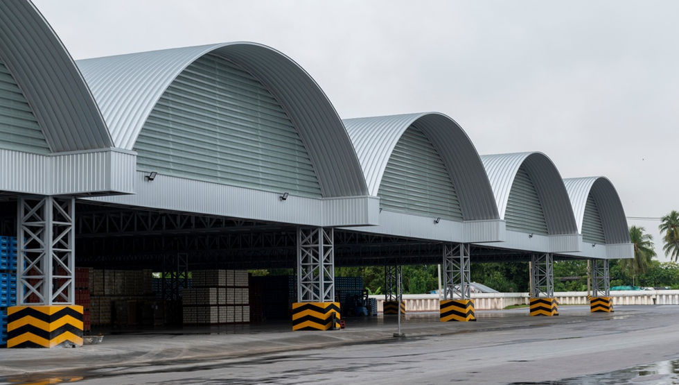 A row of large warehouse bays with curved metal roofs, striped pillars, stacked pallets, and wet pavement.