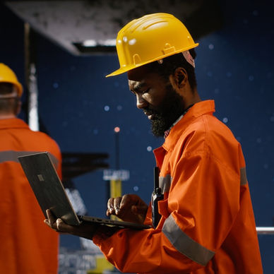 A Black man in a reflective jacket and a yellow hard hat stands, holding a laptop. There are pumps and a man behind him.
