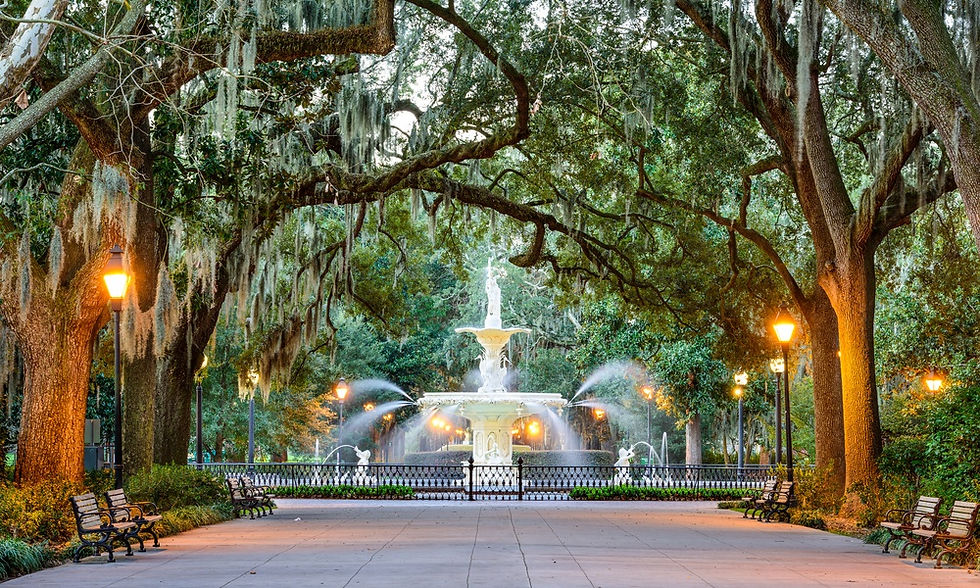 A fountain framed by moss-draped oak trees and glowing lampposts along a peaceful walkway in Forsyth Park in Savannah, GA.