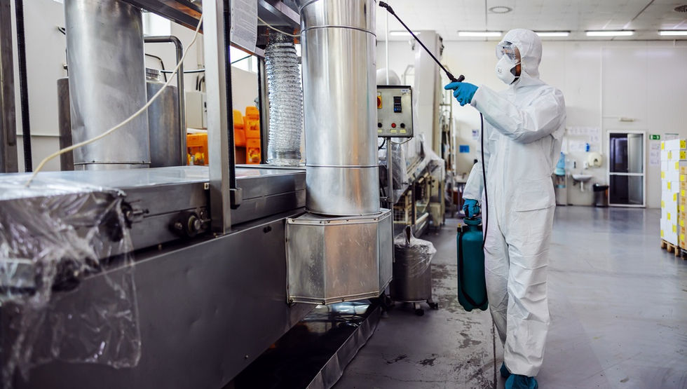 Technician in white safety coveralls uses a sprayer near large metal ducts and machinery in a clean factory.