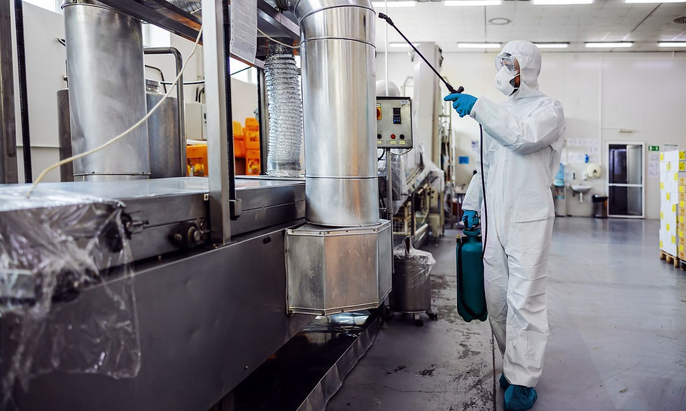Technician in white safety coveralls uses a sprayer near large metal ducts and machinery in a clean factory.