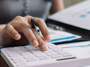 A person holding a pen presses a calculator beside printed financial charts and business reports spread across a desk.