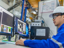 A man wearing a hardhat and safety glasses sits at a computer desk in a manufacturing facility. A program is open on the computers.