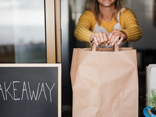 A woman in a yellow sweater with a white apron holding a paper bag out of a window next to a chalkboard that says "TAKEAWAY."