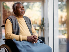A senior man sitting in a wheelchair next to a window while looking outside, with a blanket across his lap.