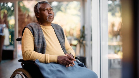 A senior man sitting in a wheelchair next to a window while looking outside, with a blanket across his lap.