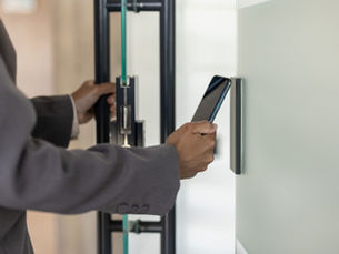 A person in a gray blazer holding a smartphone near a wall access panel beside a glass office door with a black handle.
