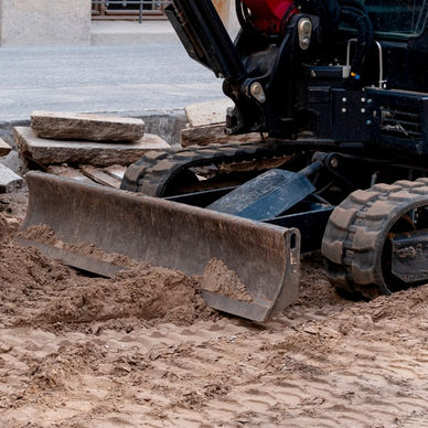 A close-up of a red track loader with a small bucket-like attachment driving over a section of dirt next to a paved area.