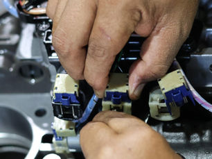 A close-up view shows a person fixing a solenoid valve. The valve is blue and cream-colored and surrounded by metal pieces.