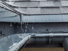 Rainwater flows from a roof into a gray gutter beneath wet shingles, with water droplets visible along the roof edge.