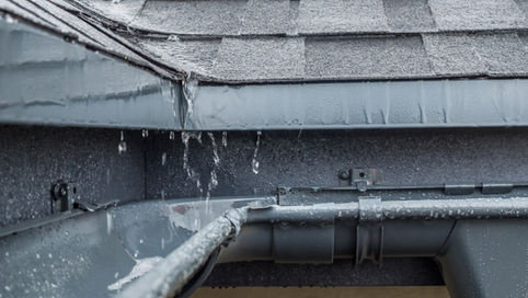 Rainwater flows from a roof into a gray gutter beneath wet shingles, with water droplets visible along the roof edge.