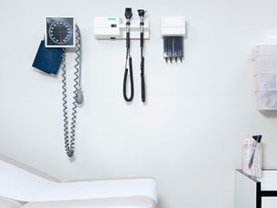 A medical exam room wall with blood pressure cuff, diagnostic tools, supply bins, cabinet, and padded exam table.