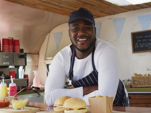 A young Black business owner smiles at the camera as he leans on the counter of his small, local food truck.