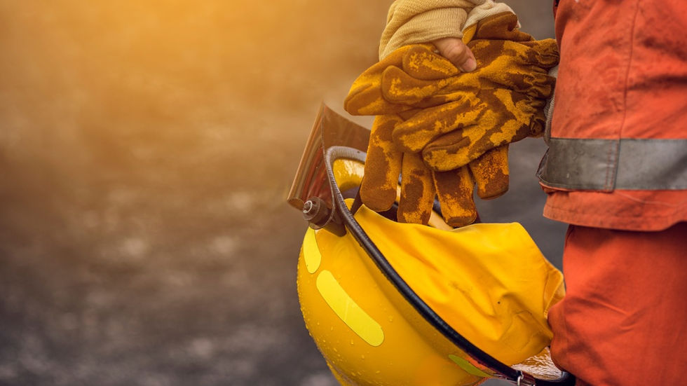 A firefighter holding a yellow helmet and protective gloves while standing near orange flames and spraying water.