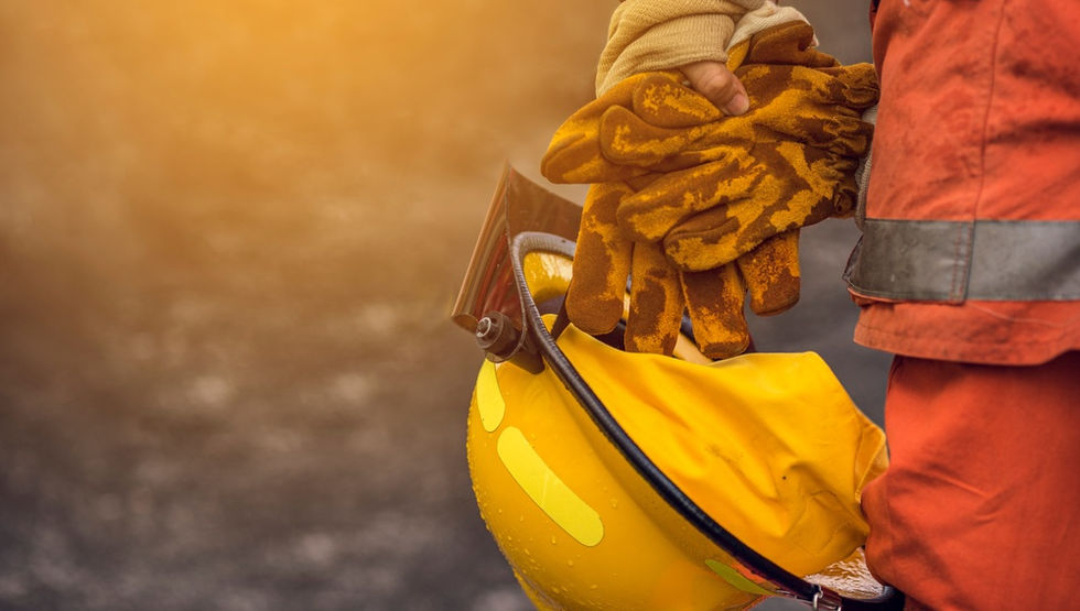 A firefighter holding a yellow helmet and protective gloves while standing near orange flames and spraying water.
