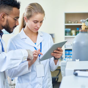 A man and a woman wearing white lab coats stand in a lab looking at a tablet next to two gas cylinders.