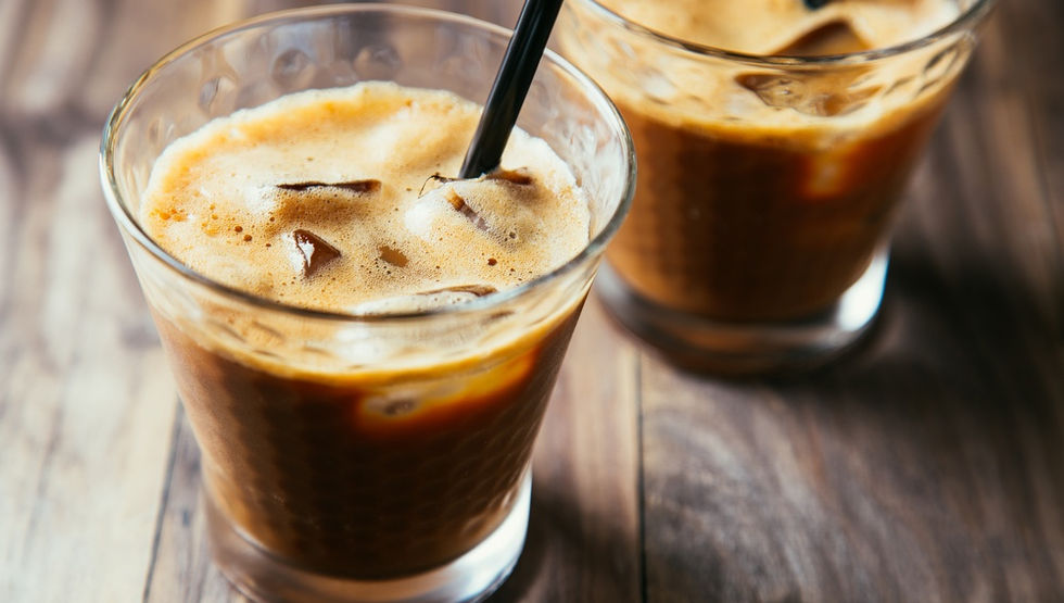 Two glasses sit on a wooden tabletop with ice and black plastic straws. The coffee beverage is a latte with foam.