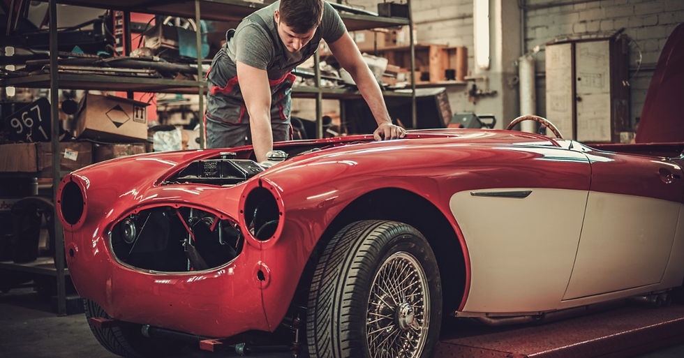 Mechanic works on a red and white classic convertible with its headlights removed inside a cluttered repair garage.