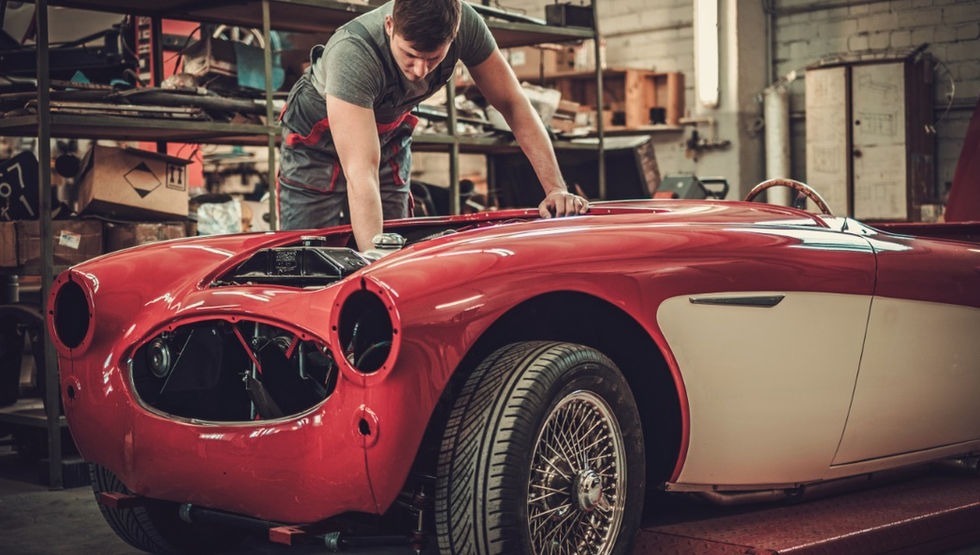 Mechanic works on a red and white classic convertible with its headlights removed inside a cluttered repair garage.