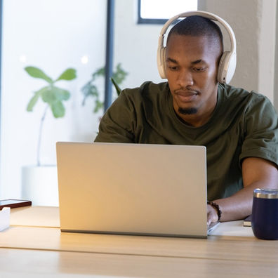 A man wearing headphones works on a laptop at a bright office desk with a lamp, coffee tumbler, and phone.