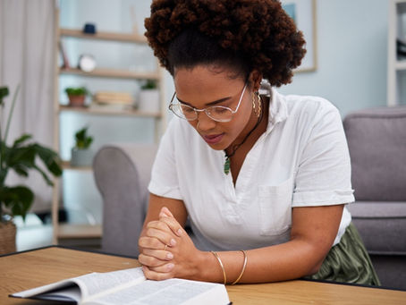 A Black woman kneeling at a coffee table inclines her head over an open Bible and clasps her hands in prayer.
