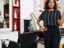 A hairstylist holding a round brush while standing beside a salon chair with mirrors and styling stations.